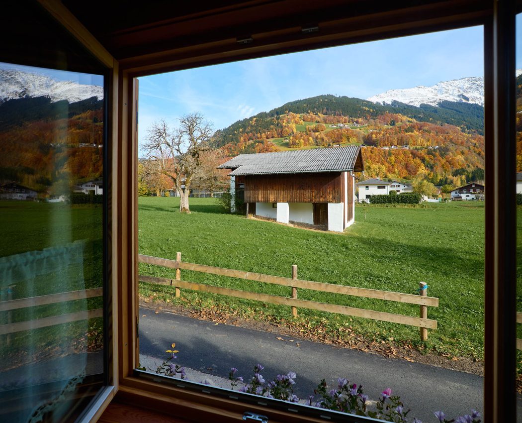 offenes Fenster mit Blick auf große Wiese mit Stallgebäude vor Bergpanorama