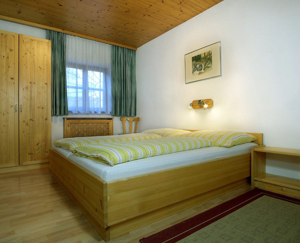 helles mit Holzmöbeln ausgestattetes Schlafzimmer. Zu sehen Doppelbett und ein Schrank neben dem Fenster