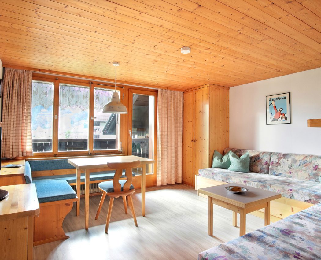 Helles Wohnzimmer mit Holzdecke. Große Fensterfront inklusive Balkontür mit Eckbank vor dem Fenster und Betten mit Couchfunktion mit Blick auf die Fenster